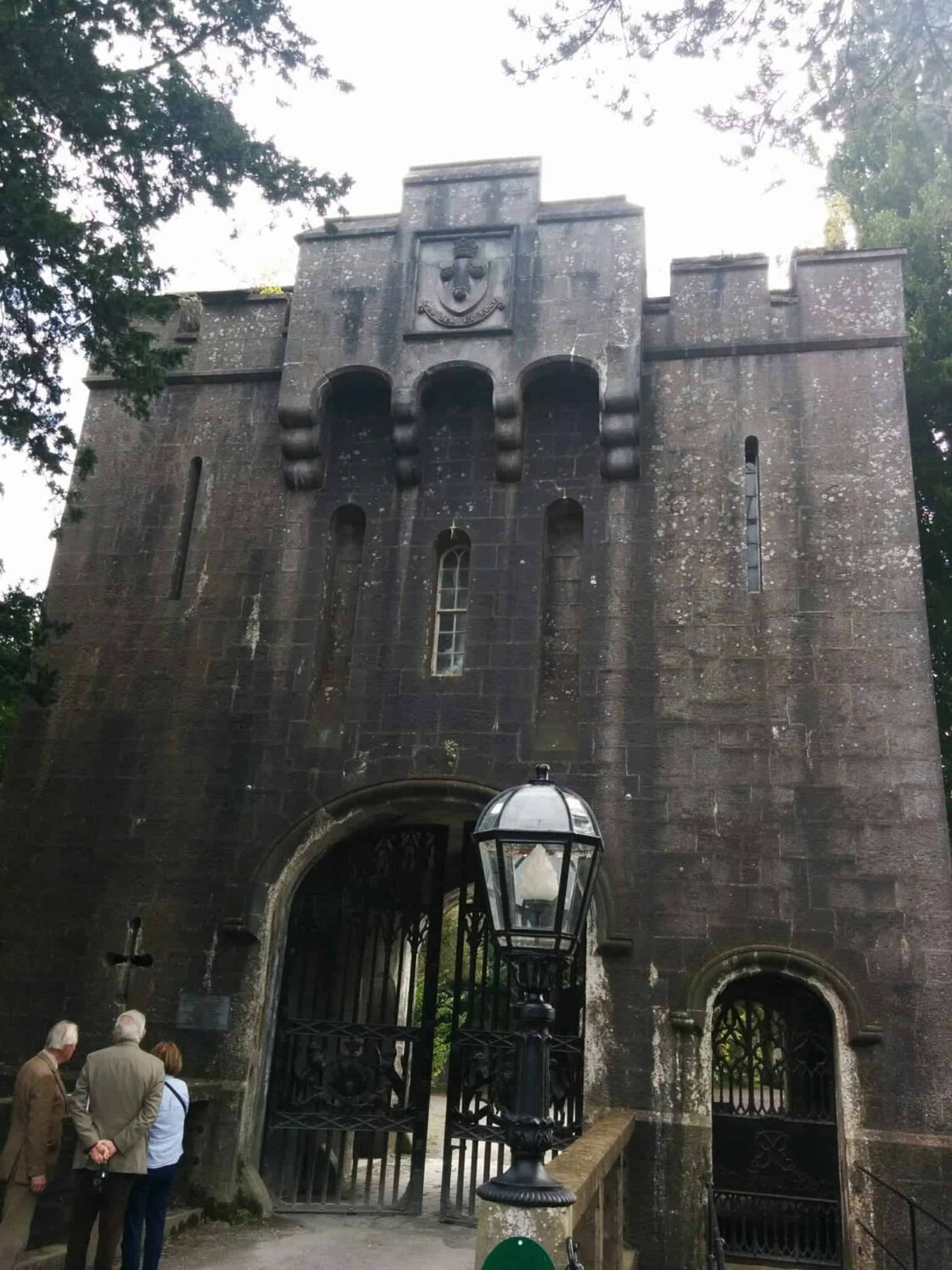 The entry Gates to Birr Castle in Ireland. The stone is a very dark grey rising up above a stone bridge over what would have been a moat centuries ago. The heavy steel gates are decorated with the family crest and the top of the gates are crenellated