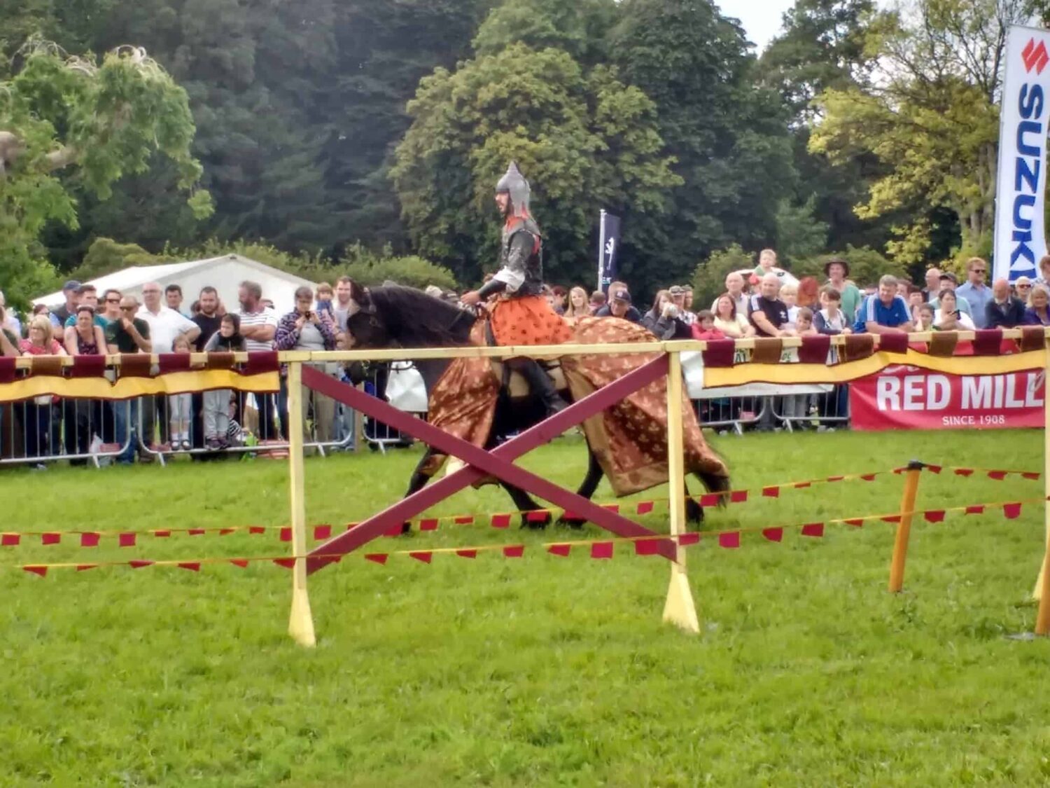 The knight is beginning his joust on a black horse he has armour and chain mail over an orange surcoat.