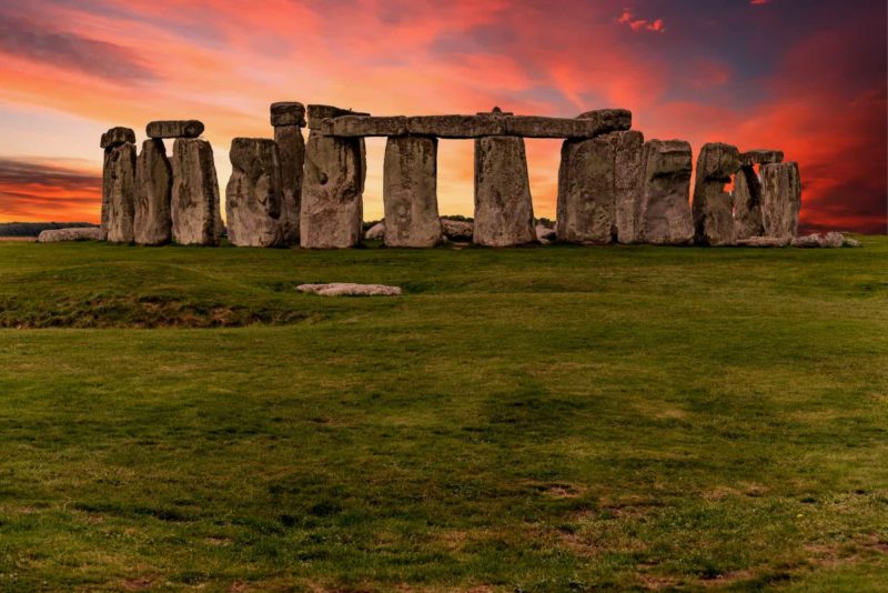 Irish folklore tells the tales of stone circles and cairns like this one.  An ancient stone henge at dusk with a pink and blue sky and a large stone circles comprised of at least 16 huge stones standing upright with large slabs laid across several of them. 