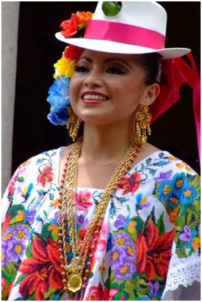 A Mexican woman poses in a white hat with a pink ribbon and flowers in her hair. On her ears are large gold earrings and she has very heavy gold necklaces on. The dress is elaborate embroidered with flowers and it is a Huipile a Mexican embroidered dress.