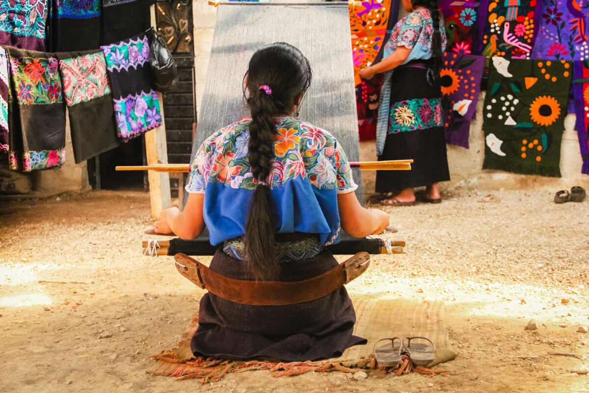 A Mexican women sits with her back to the photographer she is at a loom weaving textiles for clothing. The loom is quite long and is a lap loom. Hung either side of the loom are beautiful embroidered cloths for dresses.
