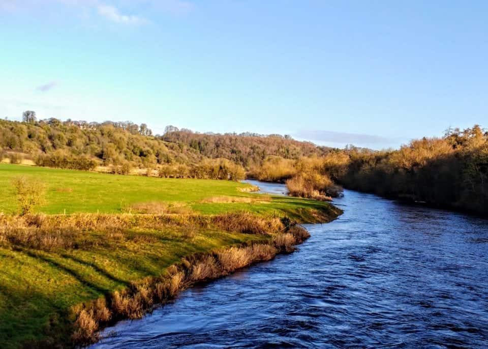 The River Boyne that runs through Meath in Ireland. The water is a very dark blue and the banks that surround it are covered in green grass, the trees are turning fall colours.
