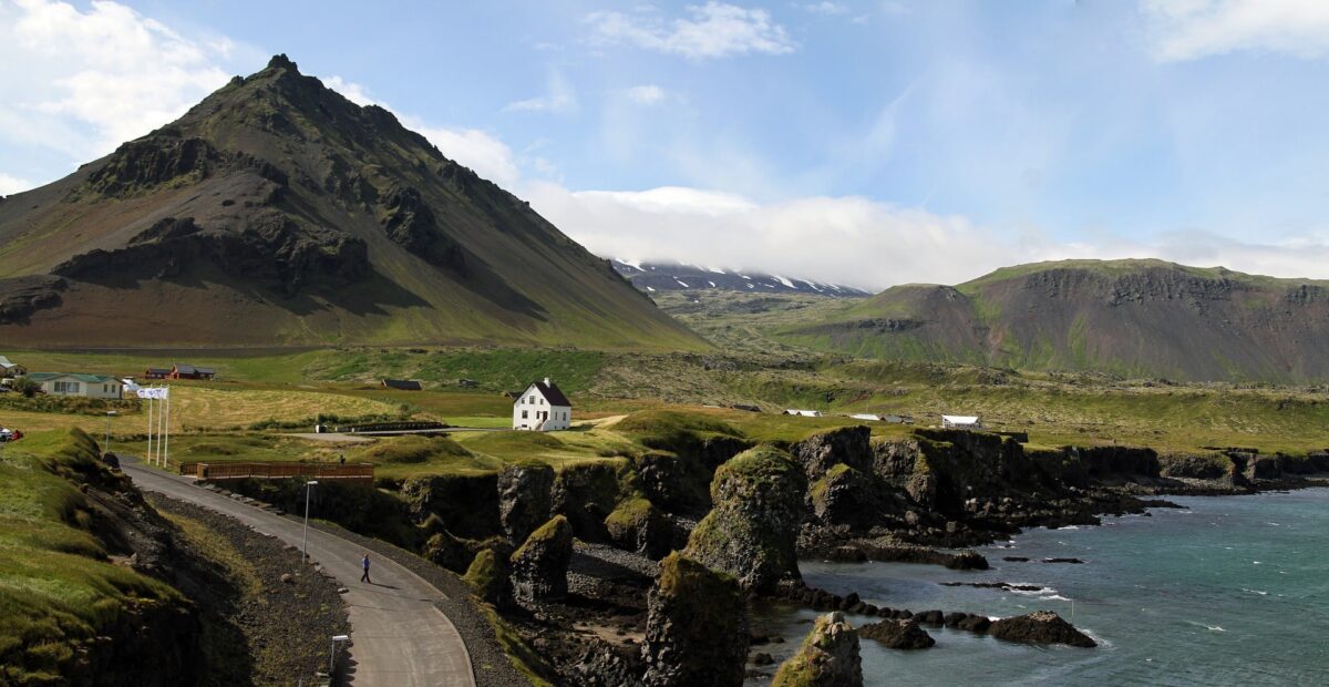 Iceland - a view of the rugged mountains and coast of Iceland. There is a gray concrete road stretching into the distance with small white houses overlooking the sea.