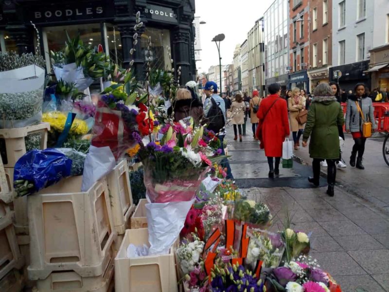 Shopping in Dublin's Grafton Street which features in many Ireland in movies. Shoppers pass by a colourful flower stall weighed down by lots of shopping bags.