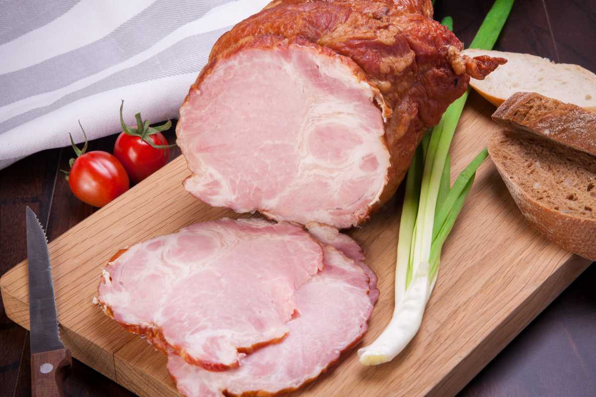 A Gammon ham sits on a board with a few slices off the meat and a decorative scallion and brown bread beside it