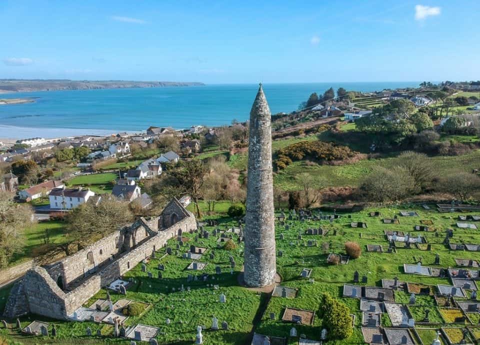 an aerial photo of the Round Tower in Waterford Ireland. The tower stands in a graveyard by a ruined church, you can see the waters of the Irish sea and the village houses that surround the ruins. One week in Ireland