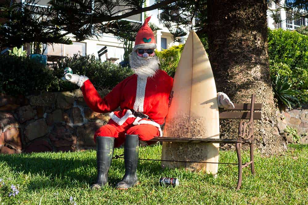 An Australian santa sitting on a bench holding a surf board in the bright sunshine