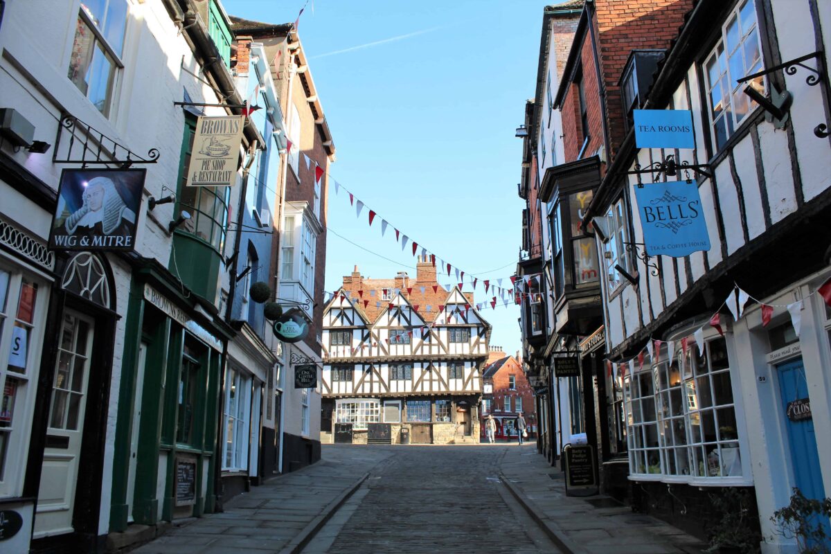 Steep Hill in Lincoln with its half-timbered medieval buildings running downhill. The buildings include a pub called the Wig & Mitre and a coffee shop with other various cafes and shops on the street. 