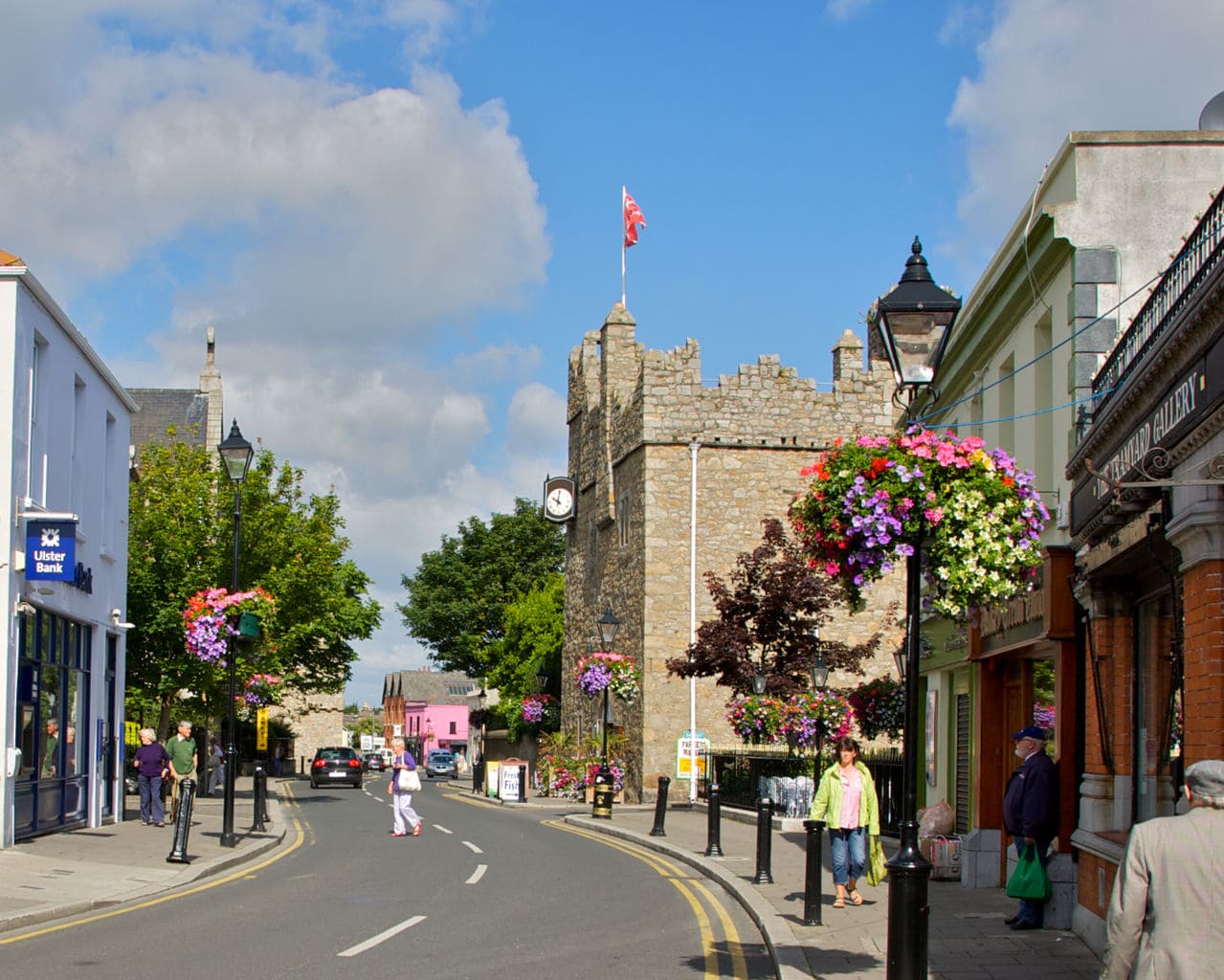 A beautiful shopping street in Howth in Ireland's Ancient East. There is a ruined castle battlement in the centre of the picture and baskets of colourful flowers hang from lightposts