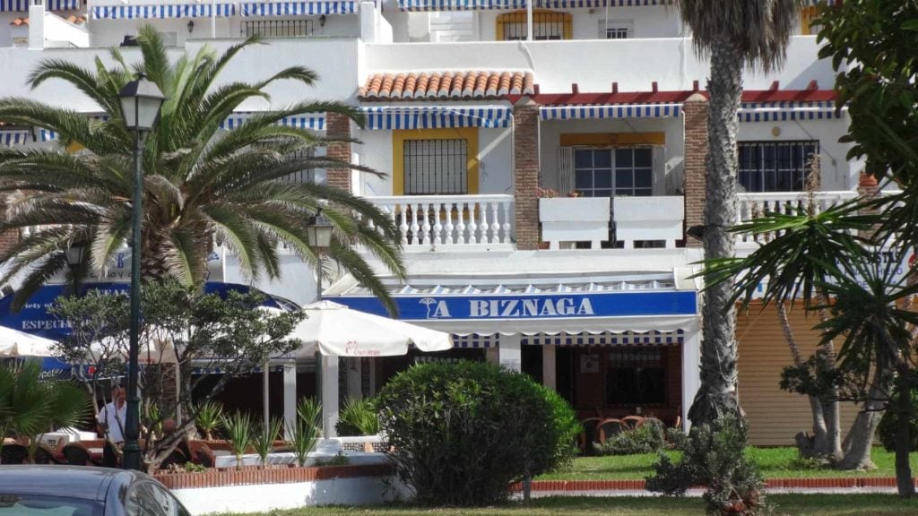 La Biznaga Salobrena Spain the exterior of a white Andalucian restaurant with striped blue awnings and a blue sign with palm trees in front and an outdoor partio with white umbrellas 