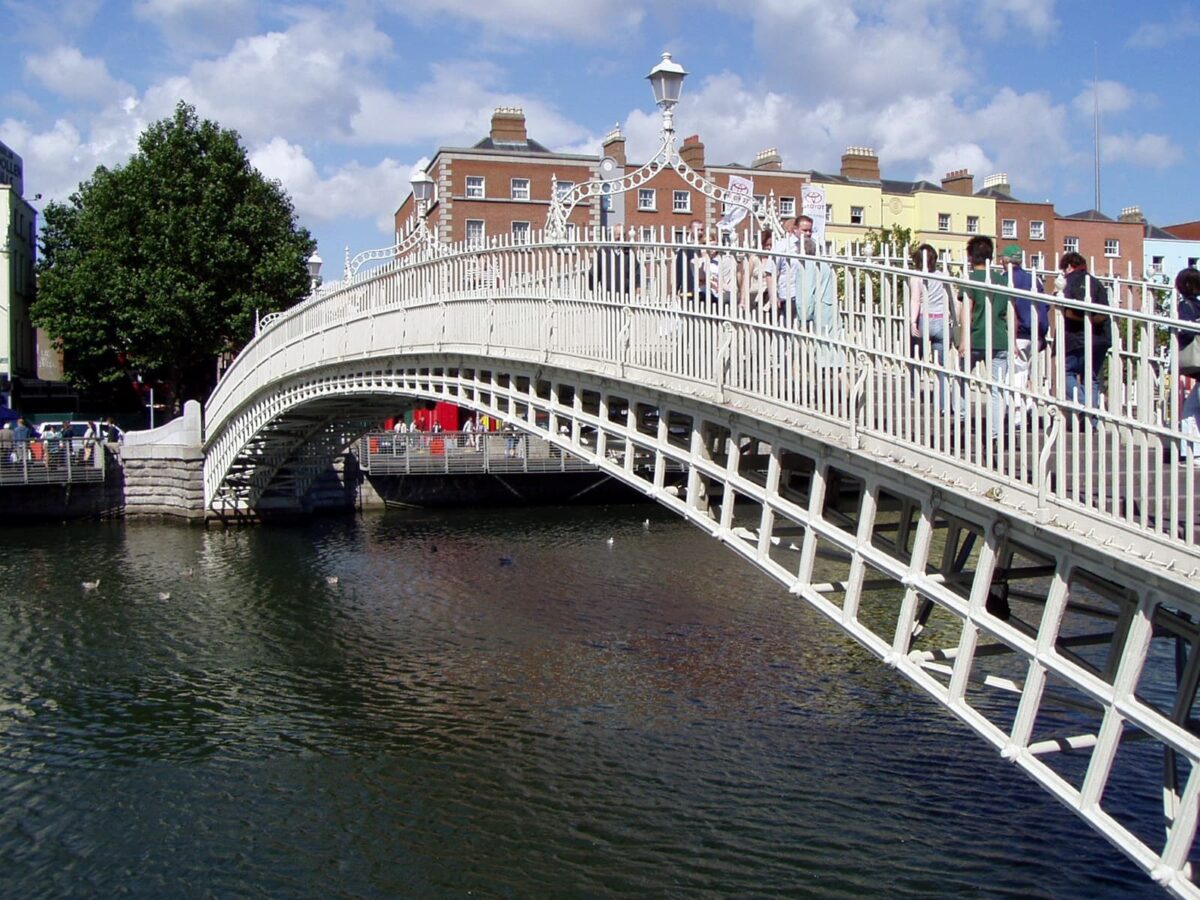 The Ha'penny Bridge in Dublin crosses the Liffey River. This famous iron bridge is all-white cast iron and is pedestrian-only. Crossing on foot are several tourists and there are ducks swimming in the river below. One week in Ireland 