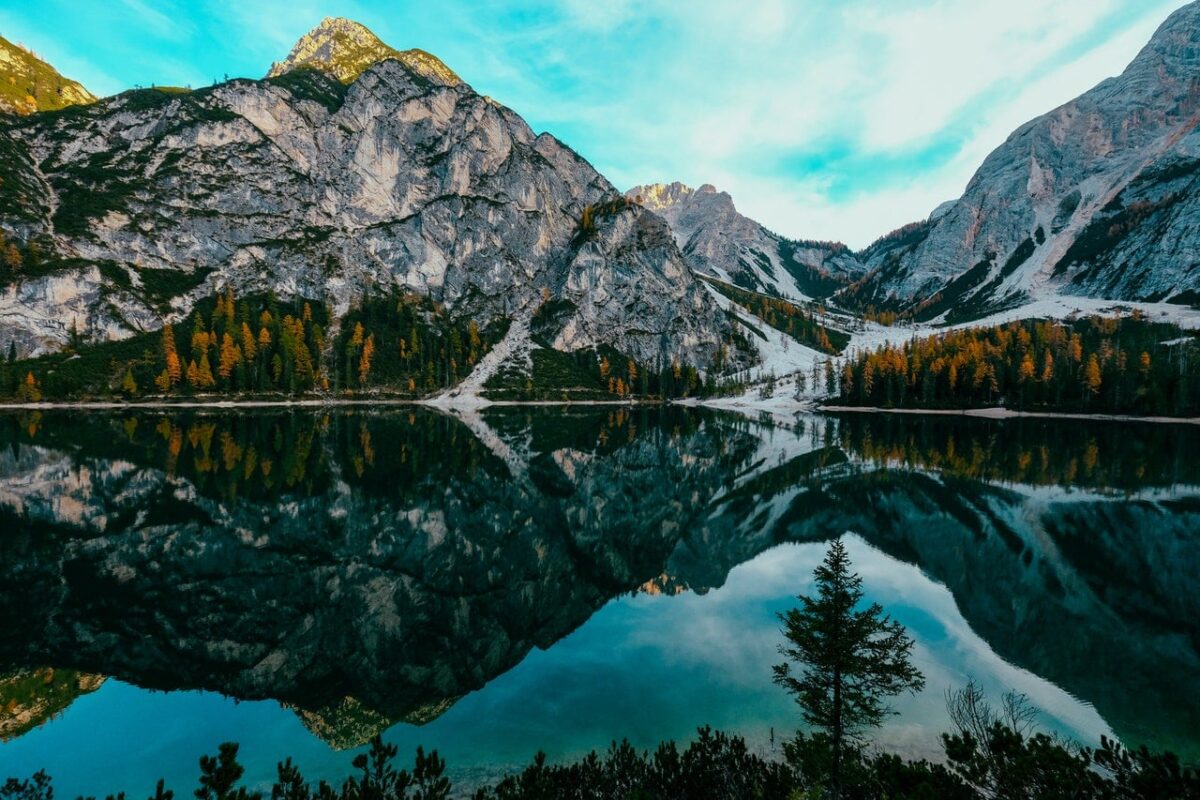 A beautiful view of the Canadian Rockies with snow craggy mountains in the background and yellow, orange and green pine trees standing around a clear lake with the mountains reflected in the lake waters 