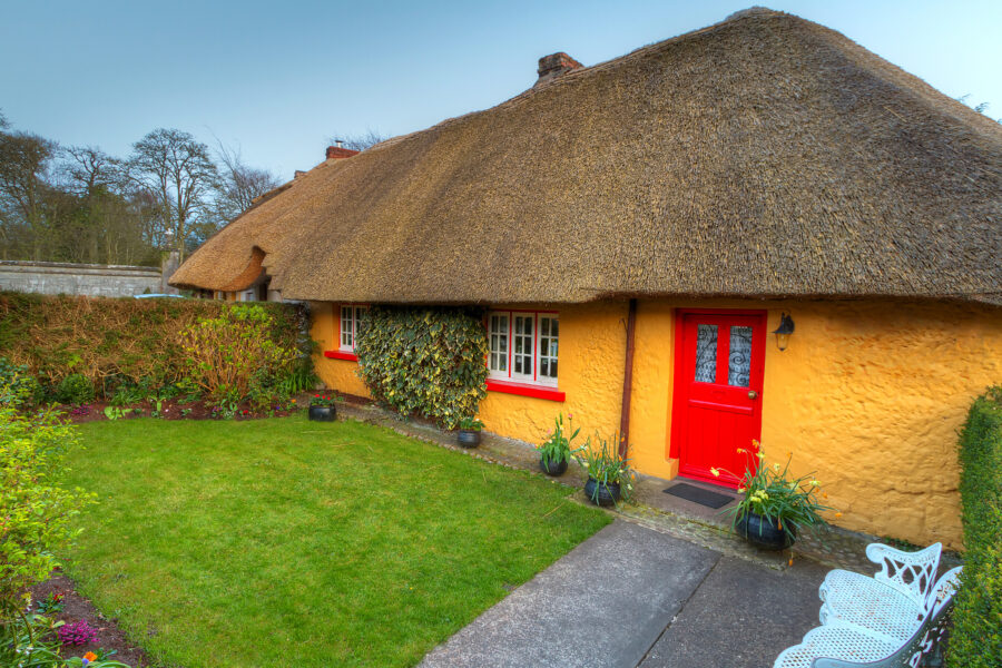 Irish Cottages to rent. Gorgeous golden thatched cottage in Ireland. The cottage has a red door and window trim and a small area of green grass in front with white cast iron seats and table