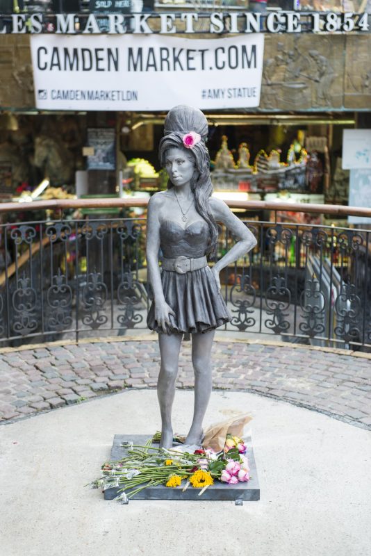 Amy Winehouse Statue in Camden Town. Silver coloured statue of Amy with flowers laid at its feet