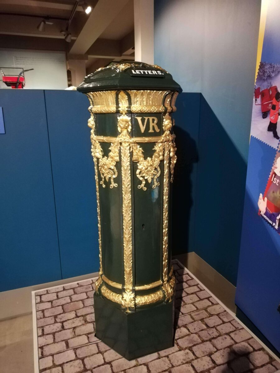 Britain's first mailbox at the British Postal Museum. It's a very dark green with lots of gold gilt dripping from the decorations on the round pillar box style with a VR which is the Royal crest