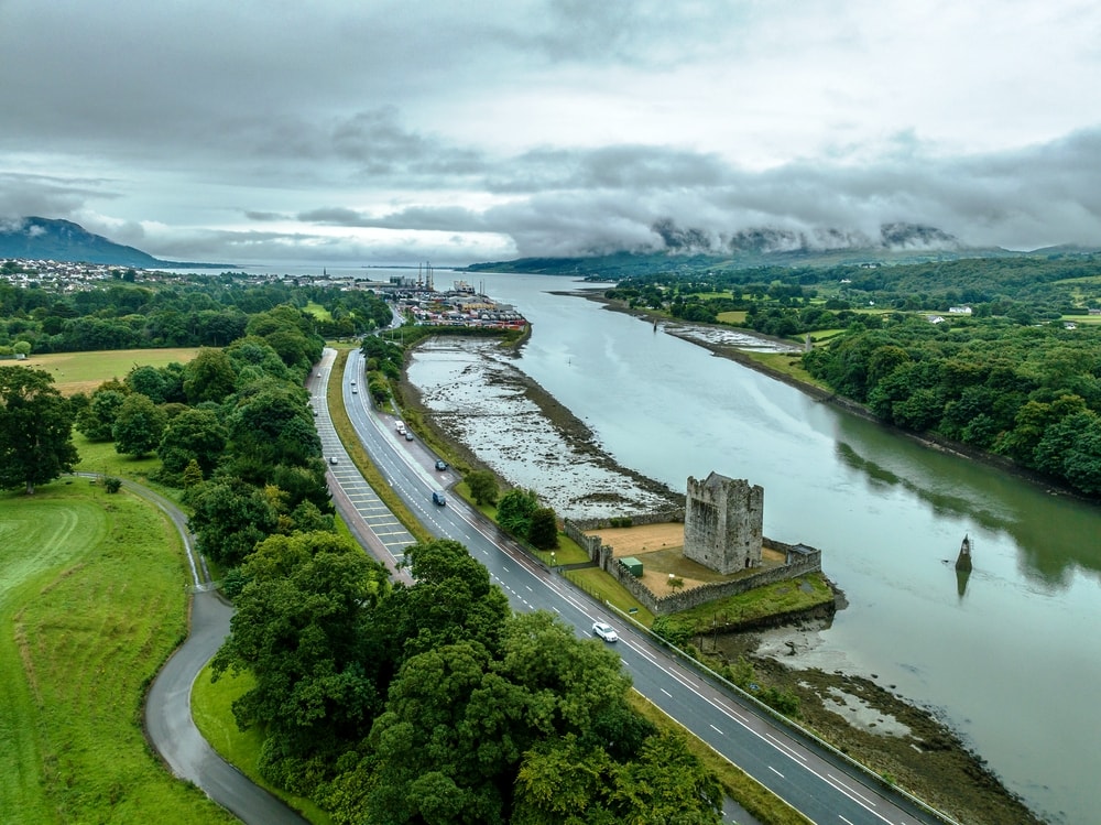 Aerial view of a river with a medieval tower north of Dublin, adjacent road, and lush greenery under a cloudy sky.