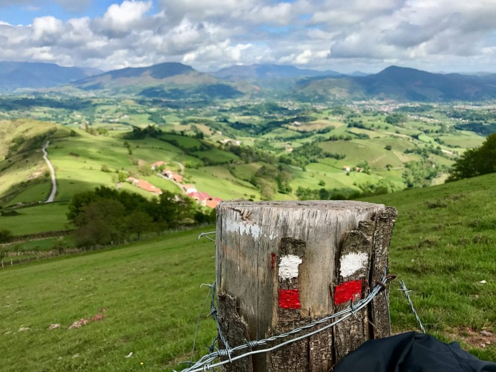 Hike the Camino a signpost overlooking a verdant green valley with small terracotta houses in the distance down in the valley and mountains in the background