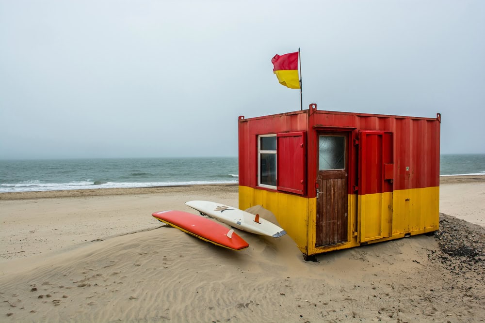 A small red and yellow lifeguard hut with a flag on the roof beside two stacked surfboards on one of the best beaches, even in the fog.