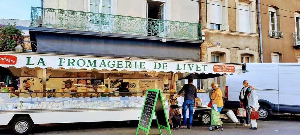 An open-air market in France with a cheese van that id about 20 feet long and has several customers lining up for the cheese.