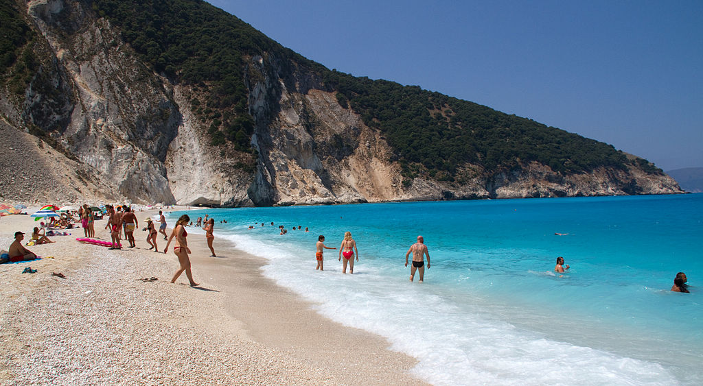 Underrated Destinations in Europe people on a gorgeous beach in Kefalonia called Mytros the beach sits at the feet of two mountains with pink sand that glows from the marble that washes up on the shores