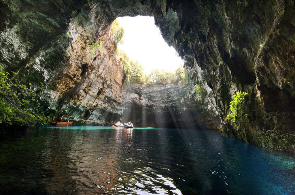  Underrated places in Europe to visit -a view of the Melissani cave in Kafalonia Greece. A cave filled with water with people in a boat and the sun streaming through a vast hole in the cave's ceiling. 