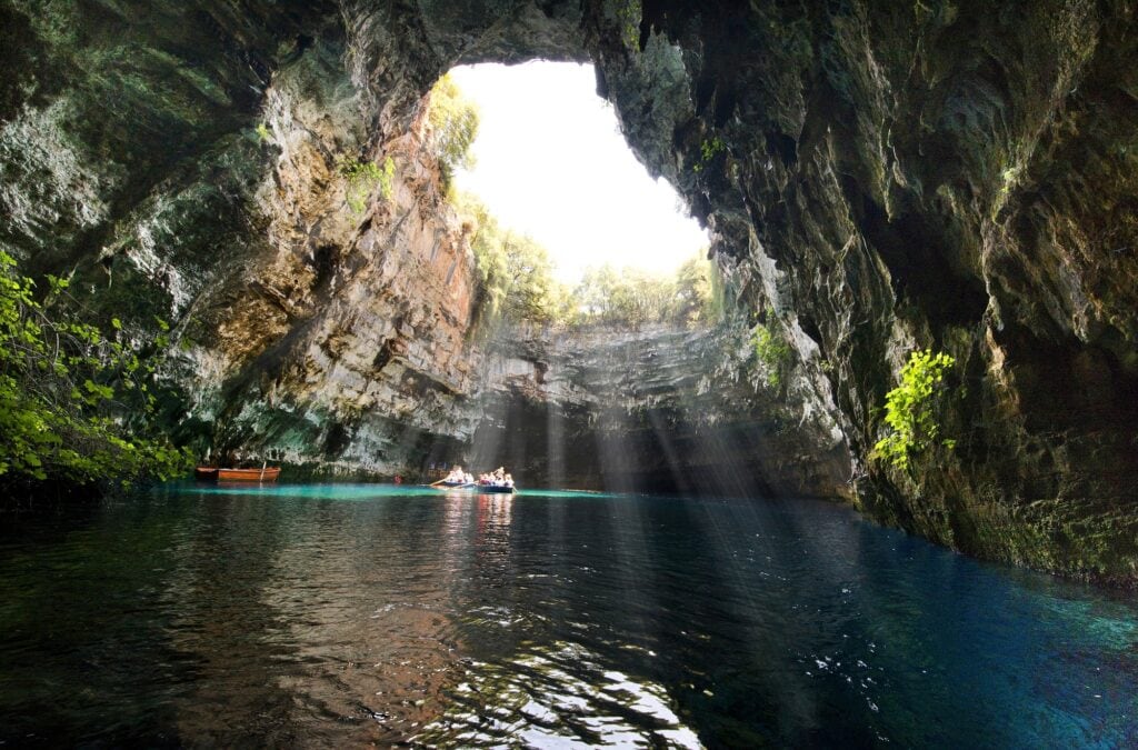 5 Underrated Destinations in Europe a view of the Melissani cave in Kafalonia Greece. A cave filled with water with people in a boat and the sun streaming through a vast hole in the cave's ceiling. 