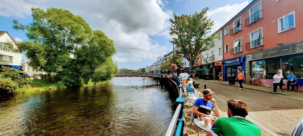 Things to do in Sligo hanging out by the river with glorious hanging flower baskets several folks are sitting at small tables enjoying a coffee.