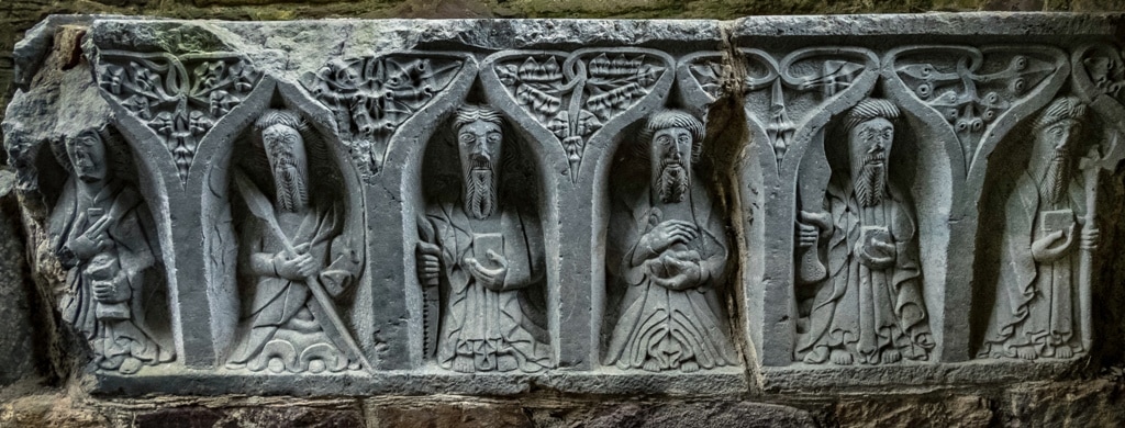 a row of famous Irish saints adoring a ruined cathedral. The images are carved into grey stone with decorative elements including each holding something important to Christianity