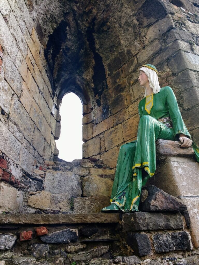A statue of a medieval woman in a green dress with gold trim and a head scarf of white linen sits in the arch of a window at Carrickfergus Castle. She is Affreca wife of John de Courcy and she founded Grey Abbey in County Down