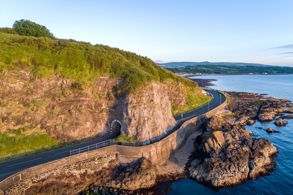 Black Arc tunnel and Causeway Coastal Route. Scenic road along eastern coast of County Antrim, Northern Ireland, UK. Aerial view in sunrise light tips for driving in Ireland.