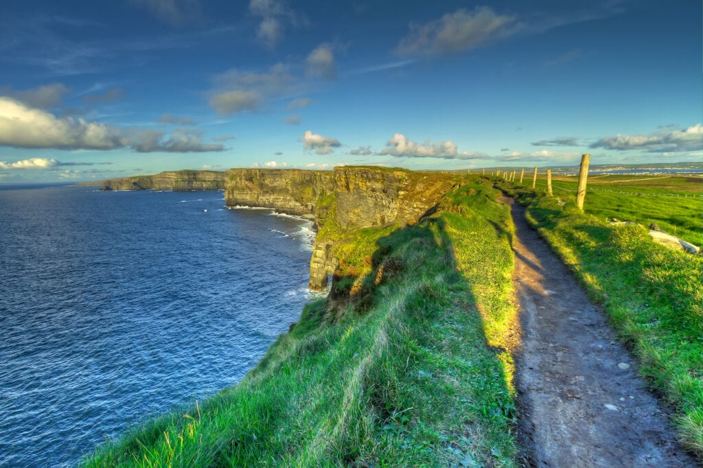 Renting a car in Ireland, a view of the Cliffs of Moher and the narrow pathways that run between the grassy verges. To the right there are fence posts that keep the cows from falling off the cliffs
