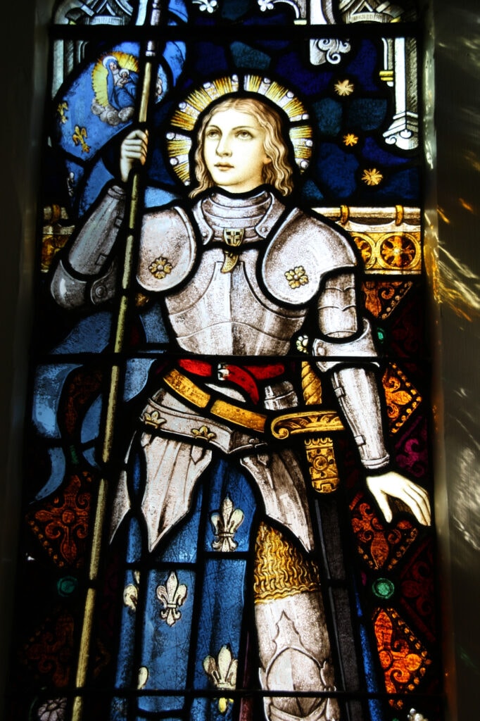 A stained glass window depicting the patron saint of France Joan of Arc. Joan appears with full silver armous and a halo above here head. The Madonna is pictured to her right just above her right hand. 