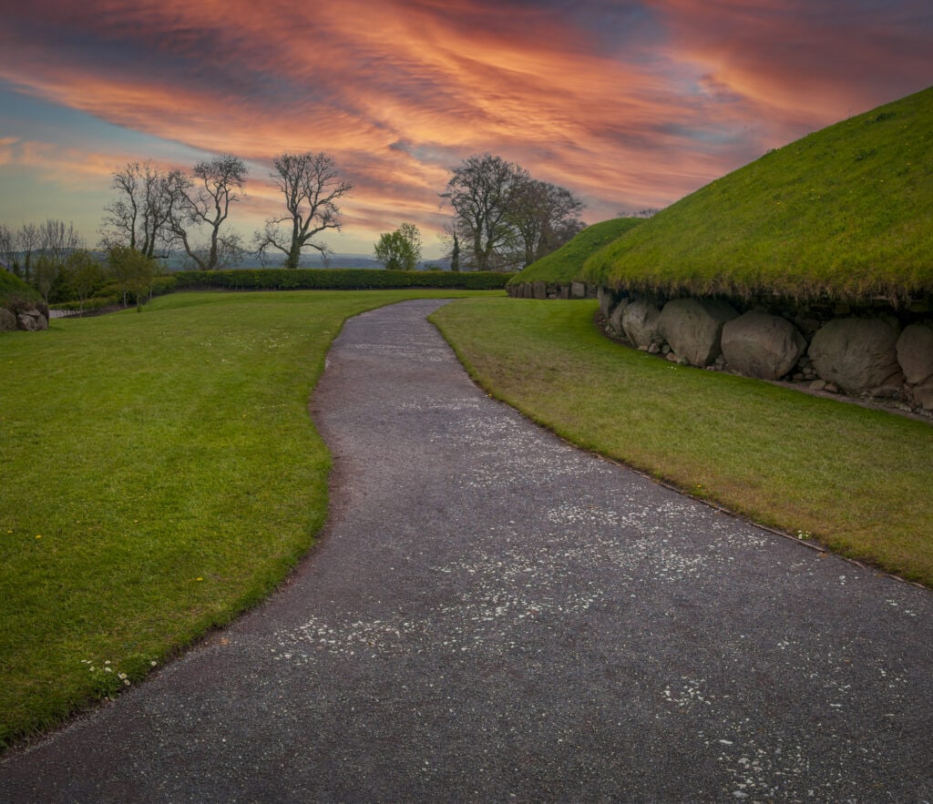 Sunset at Knowth - a neolithic tomb with large stones at the base and a green mound above it which is the tomb. There is a concrete path running around the round hill that is the tomb.