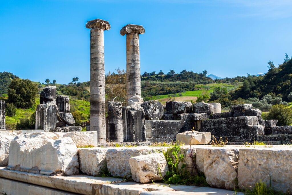 The ruins left of the Temple of Artemis at Ephesus. Two columns stand about 12 feet high with a wide variety of time-worn blocks of granite at their feat.
