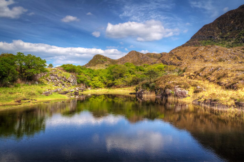 Ladies view lake a calm blue reflects the mountains and hills of the Ring of Kerry. Several shades of green surround the lake and the skies are reflected in the lake