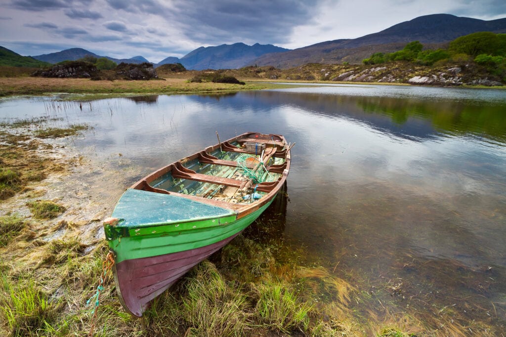 An old rowboat sits on the lake it is green and purple and quite weathered. There are the Killarney mountains in the background and the lake is a calm deep blue