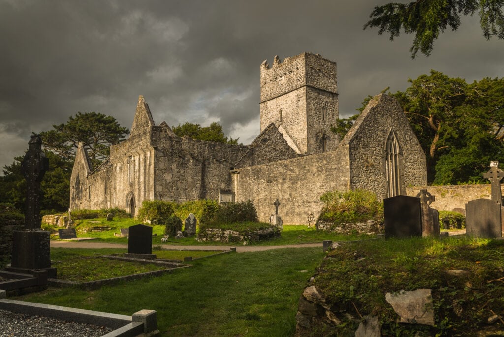 The ruins of Muckross Abbey with its stone walls. There is no roof and the ruins are surrounded by an old graveyard with tip tilty head stones and small Irish crosses