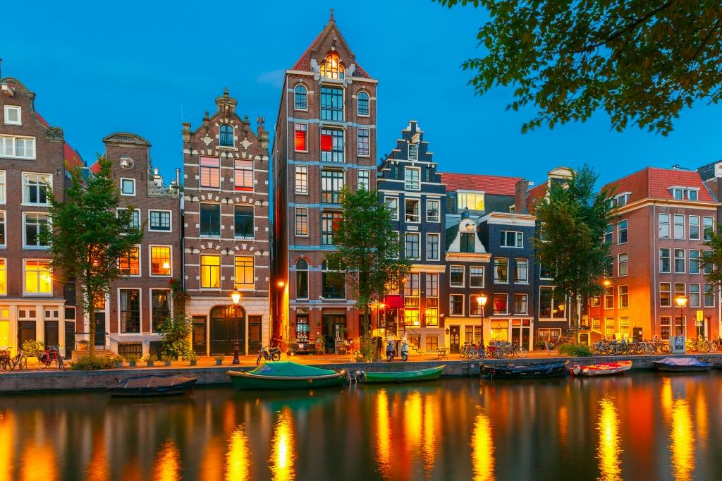 A view of houses in Amsterdam when the sun has just set and there is a golden glow over the canal. The houses are all row houses and in typical Dutch style with pointed roofs and lots of windows.