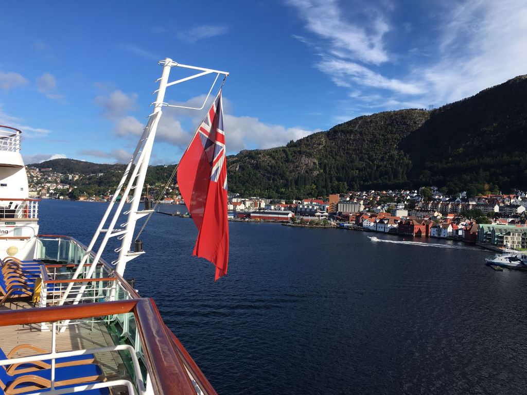 View of the coast of Norway on a Norwegian Fjords Cruise. The flag flies from the bow of the ship with views of a Norwegian village and mountains in the back ground.