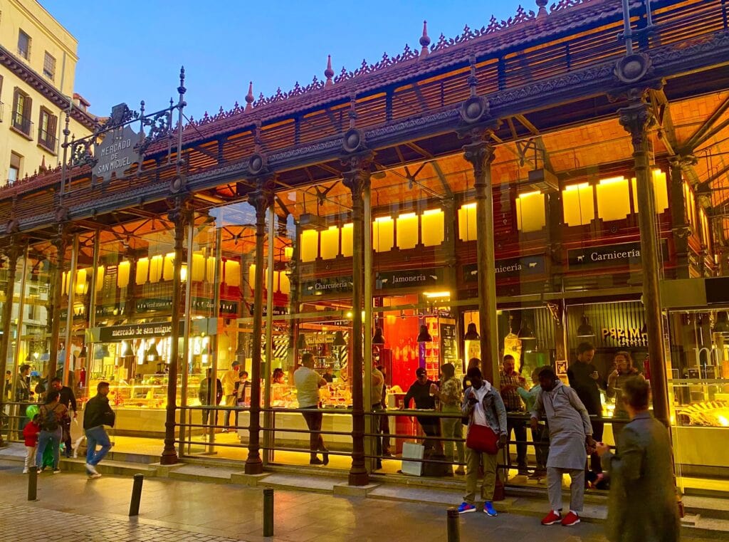 Twilight over a bustling outdoor market in Seville with illuminated stalls showcasing Spanish food and eager shoppers.