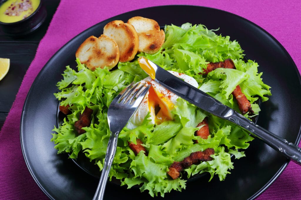 frisee lettuce, crispy bacon and a poached egg salad  -  classic Lyonnaise Salad, served on black plate with toasted baguette, authentic recipe, view from above, close-up