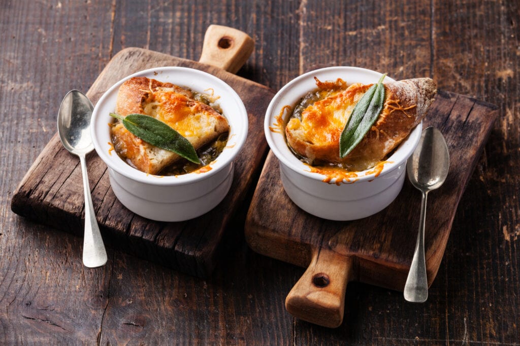 Two bowls of french onion soup sit on a wooden server with spoons. The soup is in a white bowl and you can see a french baguette on the bowl topped with a sage leaf and melted cheese.