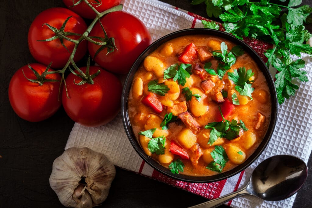 White bean soup with potatoes, tomatoes, paprika, and bacon in black ceramic bowl. Top view