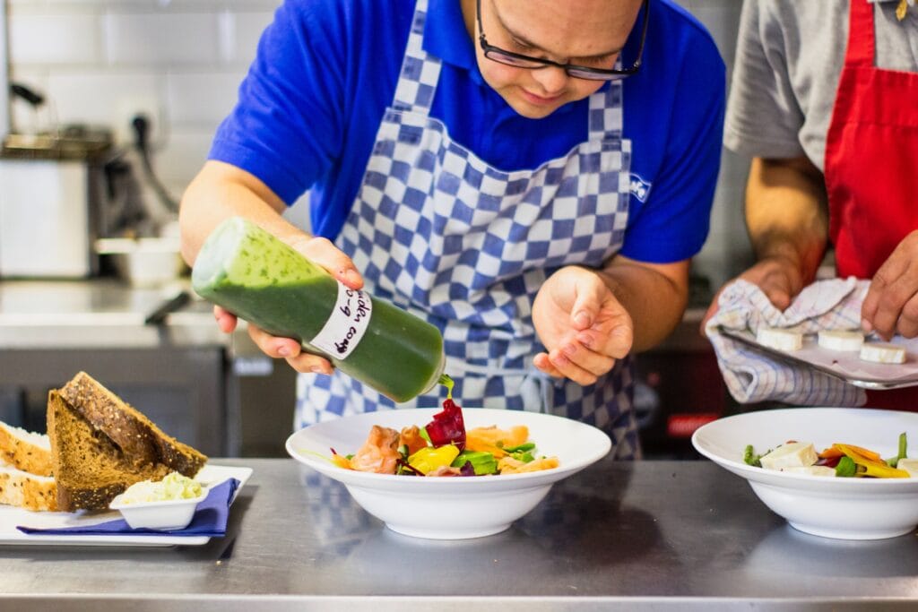 A man in a blue checked apron is using a squeeze bottle to dress a salad with a green pesto salad dressing which is in a white bowl. Cooking classes around the world