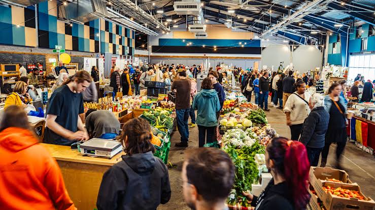 People shop at stalls filled with fresh produce inside a large, high-ceilinged indoor market hall.