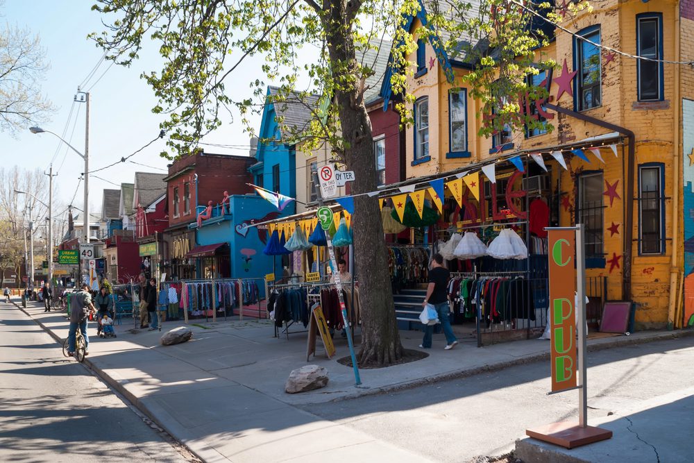 A street scene shows colorful buildings with outdoor clothing racks and decorative bunting under a tree on a sunny day. Canada travel guide
