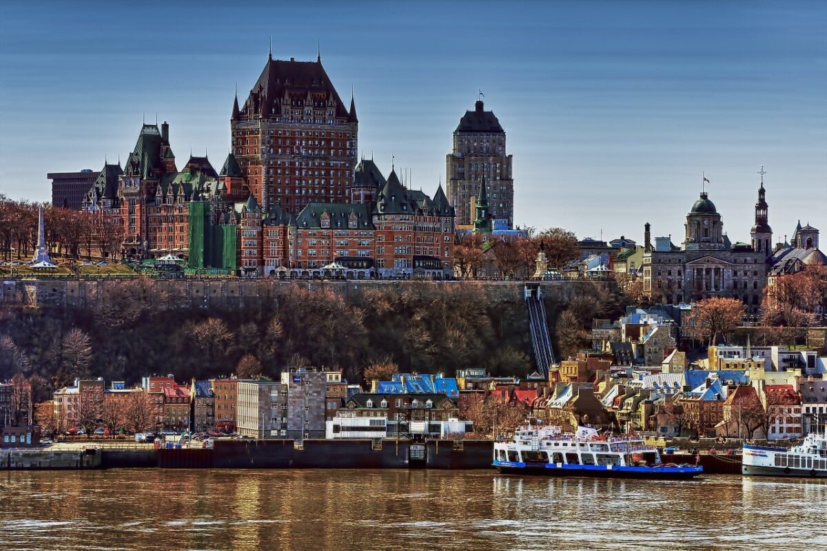 A large historic hotel sits atop a cliff overlooking a river with a ferry docked at the shore.