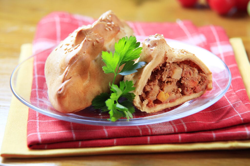 Cornish pasty history, a cornish pasty on a plate with the pasty cut open and showing the interior filled with meat, potatoes and turnip. The plate sits on a pink and white checked napkin and a clear plate