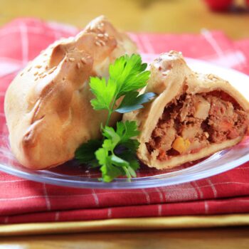 Cornish pasty history, a cornish pasty on a plate with the pasty cut open and showing the interior filled with meat, potatoes and turnip. The plate sits on a pink and white checked napkin and a clear plate