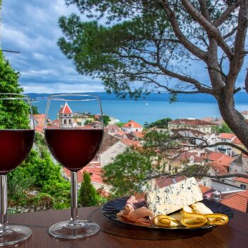 Two glasses of red wine with charcuterie assortment with view of Croatian town Omis with red roofs and blue sea. Glass of red wine with different snacks - plate with ham, sliced, blue cheese.